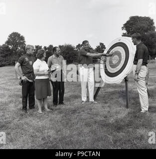 1960, historische, bei einem Dorffest eine Bogenschießen Wettbewerb, Menschen in einem Bereich, der von einem Impulsstück mit Pfeilen im Stehen. Stockfoto