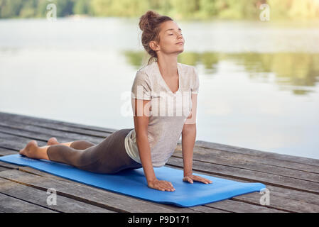 Attraktive junge Frau, auf Matte auf einem Holzdeck über einem See oder Fluss, ein Yoga mit den Augen stellen geschlossen und eine heitere Ausdruck Stockfoto