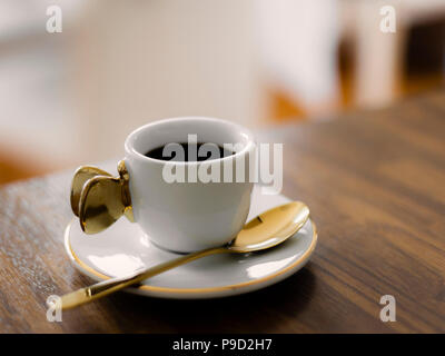 Weiße Tasse mit einem goldenen Griff, Untertasse und Löffel, mit schwarzen Kaffee auf einer hölzernen Zähler gefüllt. Stockfoto