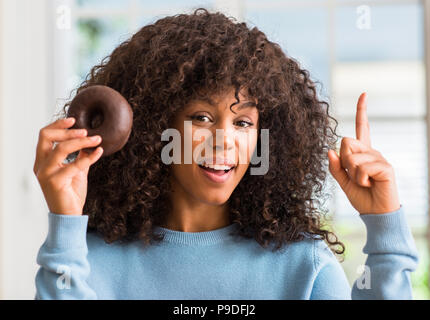 Junge afrikanische amerikanische Frau mit Schokolade Donut zu Hause mit einer Idee oder Frage Zeigefinger mit glücklichen Gesicht überrascht, Nummer eins Stockfoto