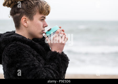 Porträt der jungen Frau mit braunem Haar und dreadlocks und eine Lippe Piercing mit schwarzen Fell Jacke, stehen, Ozean, die Tasse zu ihren Lippen. Stockfoto