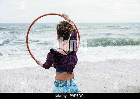 Junge Frau mit braunen Haaren und Dreadlocks, stehend auf einem Sandstrand am Meer, Balancing Hula Hoop. Stockfoto