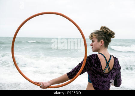 Junge Frau mit braunen Haaren und Dreadlocks, stehend auf einem Sandstrand am Meer, Balancing Hula Hoop. Stockfoto