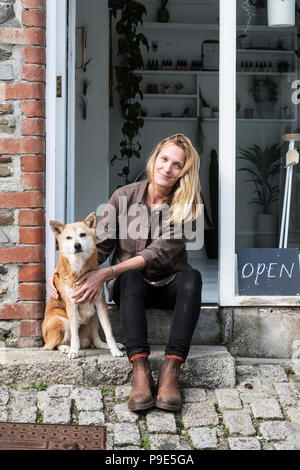 Lächelnd weibliche Besitzer von Pflanzen shop sitzen auf Schritte außerhalb ihr Speicher, ein Hund neben ihr sitzt. Stockfoto