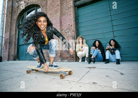 Drei junge Frauen mit lockigem Haar vor Shutter hocken, lächelnden jungen Frau, ein Skateboard. Stockfoto