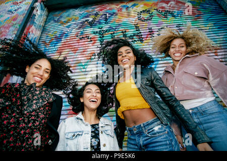 Low Angle View von vier jungen Frauen mit lockigem Haar stand vor der Blende in bunten Graffiti bedeckt, in die Kamera lächeln. Stockfoto