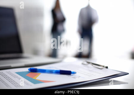 Laptop mit Ordner auf dem Schreibtisch, zwei Geschäftsleute stehen im Hintergrund Stockfoto