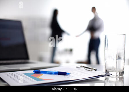 Laptop mit Ordner auf dem Schreibtisch, zwei Geschäftsleute stehen im Hintergrund Stockfoto