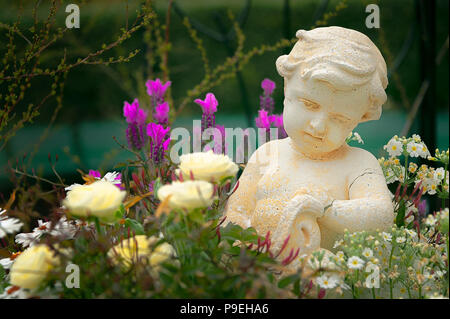 Kleine Engel in einem BLUMENGARTEN. Skulptur eines niedlichen kleinen Engel in einem Garten mit Sortiment an blühenden Sorten. Stockfoto