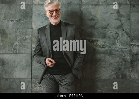 Portrait von gut aussehenden reifen Geschäftsmann ständigen entspannt auf graue Wand. Lächelnd älterer Geschäftsmann an der Kamera schaut. Stockfoto