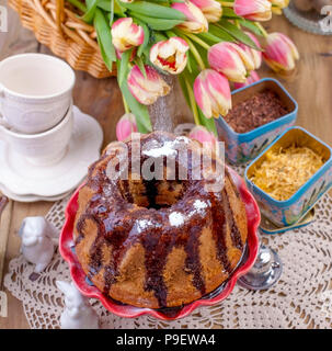 Hausgemachte süße Kuchen, gefüllt mit Schokolade, auf einem roten Kuchen Platte. Auf einer hölzernen Hintergrund, trockene Kräuter für Tee- und Cups. Einen Strauß Frühlingsblumen. Tulpen und einem keramischen Kaninchen. Postkarte und freier Speicherplatz Stockfoto
