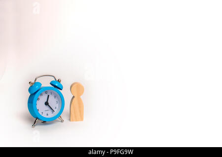 Holz- menschliche Figur steht in der Nähe ein blaues Wecker auf einem weißen Hintergrund. Der Begriff der Hoffnung und Erwartung. Warteschlange. Planung. Business, Minimalismus. Stockfoto