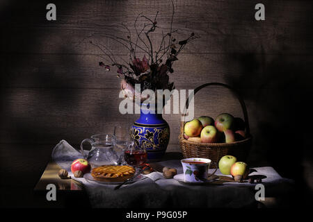 Vintage Landschaft still life: Blätter im Herbst, Ton Vase, Korb mit Äpfeln, Torte, silber Messer, antike Tee Tasse, Becher, Glas Wein, Haselnüsse, Walnüsse, Kastanien auf einem Tisch in Licht und Schatten. Stockfoto