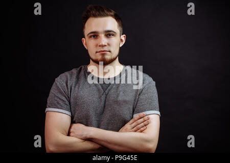Portrait von attraktiven jungen Mann in grauen t-shirt Stand mit verschränkten Armen auf schwarzem Hintergrund isoliert. Stockfoto