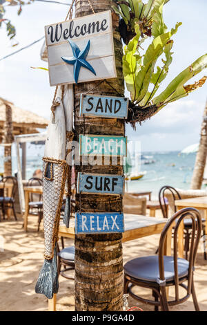 Holz- schilder Willkommen, Sand, Strand, surfen, entspannen und eine hölzerne Fisch auf einem Baum an einem Strand Cafe am Pantai Sanur, Bali, Indonesien. Bild vertikal. Stockfoto