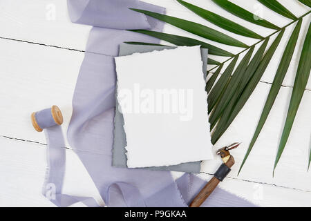 Weiße leere Grußkarte Band auf dem Hintergrund der blauen Stoff mit Palm Leaf und kalligraphischen Stift auf einem weißen Hintergrund. Mockup mit Umschlag und eine leere Karte. Flach. Ansicht von oben Stockfoto