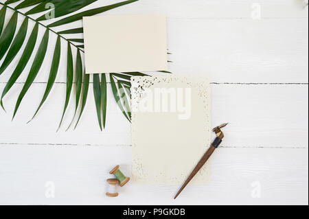 Weiße leere Grußkarte Band auf einem Hintergrund mit Palm Leaf und kalligraphischen Stift auf ein weißes Holz- Hintergrund. Mockup mit Umschlag und eine leere Karte. Flach. Ansicht von oben Stockfoto