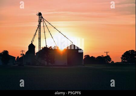 Burlington, Illinois, USA. Ein farbenfroher Sonnenuntergang fällt hinter Kooperative voller Lagerung der Ernte Tanks und Silos ein Landwirt. Stockfoto