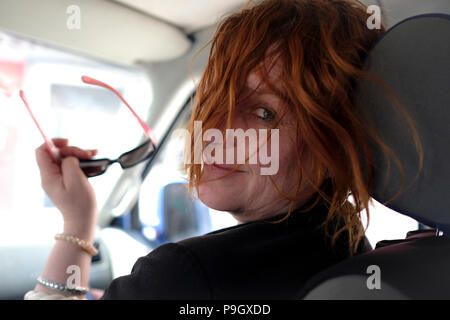 Attraktive französische Frau mittleren Alters lächelnde Blicke zurück über die Schulter im Auto mit Sonnenbrille in Ihrer Hand Stockfoto