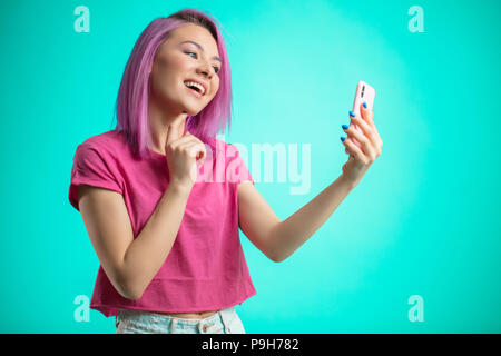 Positive lächelnde Schüler Mädchen mit rosa Haaren, in rosa T-Shirt gekleidet, so dass selfie Foto nach Friseur auf dem Smartphone isoliert auf einem blauen backgrou Stockfoto