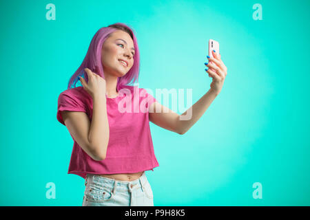 Brustbild mit einem lächelnden Frau mit rosa Haaren, in rosa T-Shirt gekleidet, so dass selfie Foto auf dem Smartphone auf einem blauen Hintergrund mit Stockfoto