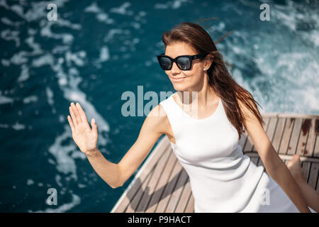 Happy kaukasischen weibliche model in weißem Kleid tragen Sonnenbrillen gruss Kapitän auf einem luxuriösen Schiff an sonnigen Morgen. Frau Schönheit, Meer Reisen Stockfoto