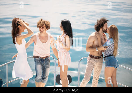 Gruppe der kaukasischen Jungs und ihre Freundinnen stehend mit erhobenen Händen auf dem elge von Yacht s Deck, glücklich mit Sonne, Meer, Wind und Urlaub. Die c Stockfoto