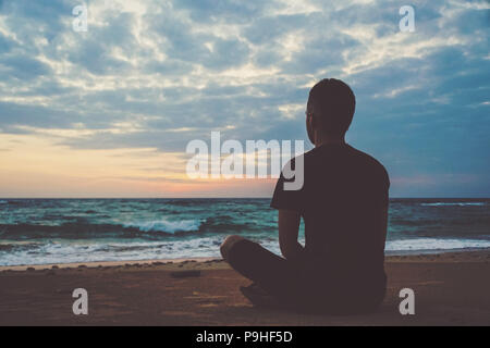 Junge Menschen meditieren auf ocean Cliff während des Sonnenuntergangs. Kerl sitzt in einem Lotus Position auf Sand am Strand bewundert und Morgendämmerung auf dem Hintergrund von Clo Stockfoto