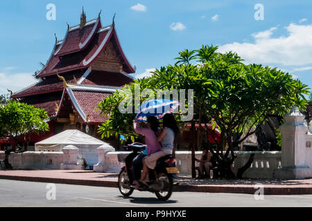 Zwei laotische Mädchen fahren mit dem Roller vor dem Wat Mai Suwannaphumaham Tempel in Luang Prabang, Laos, Holding einen Regenschirm zu schützen sich Stockfoto