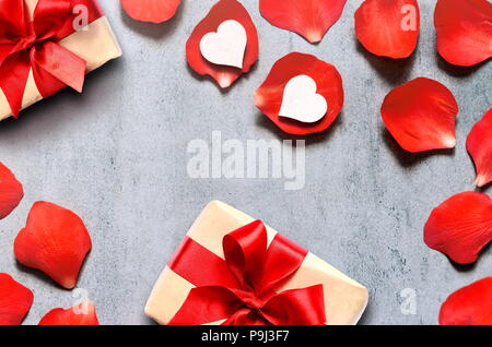 Geschenk im Handwerk Papier mit Herzen Dekoration auf roten Rosenblättern Hintergrund auf grau Tabelle. Romantik Konzept. Flach, Ansicht von oben. Stockfoto