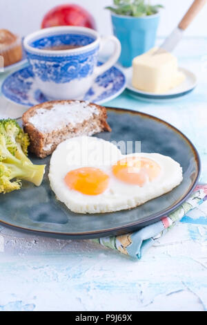 Spiegeleier in der Form von Herzen. Auf einem dunklen Platte mit Brokkoli und Toast. Blaue Tasse mit Tee, Butter zum Frühstück und Muffins. Auf einem hellen Hintergrund. Freier Platz für Text oder Werbung Stockfoto