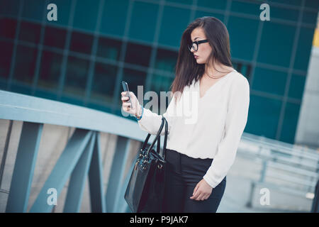 Schöne brünette Frau mit weißem Hemd von SMS-Nachrichten auf dem Smartphone zu Fuß auf der Straße in einem bewölkten Tag. Stockfoto