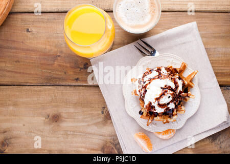 Süße Waffeln zum Frühstück, mit weißer Sahne und Schokoladensauce eingerichtet. Ein Glas mit schwarzem Kaffee und Orangensaft, auf braunem Holz- Hintergrund. Stockfoto
