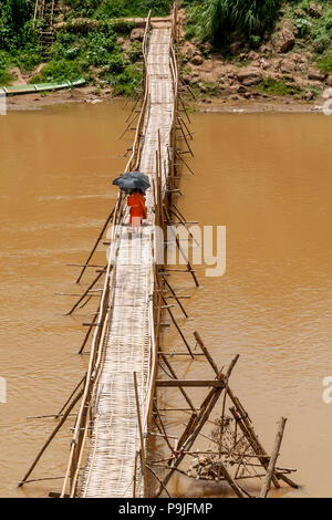 Buddhistische Mönche überqueren Sie den Fluss Nam Khan auf einem Bambus-Brücke, die sich zum Schutz vor der Sonne mit Sonnenschirmen in Luang Prabang, Laos Stockfoto