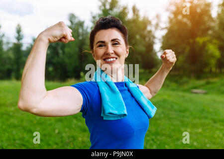 Lächelnd Senior Frau flexing Muskeln Outdoor im Park. Ältere Frauen zeigen Bizeps. Gesunden Lebensstil Konzept. Copyspace. Stockfoto