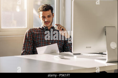 Mann, der an seinem Schreibtisch im Büro an einem Geschäft Papier suchen zu sitzen. Unternehmer lesen ein Papier sitzt vor einem Computer im Büro. Stockfoto