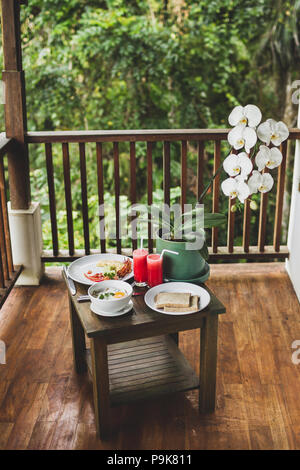 Frühstück im Freien auf der Terrasse aus Holz mit Jungle View. Eier, frische Wassermelone Saft und Orchideenblüte auf Tisch Stockfoto