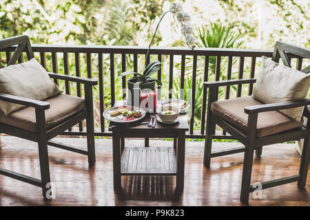 Frühstück im Freien auf der Terrasse aus Holz mit Jungle View. Eier, frische Wassermelone Saft und Orchideenblüte auf Tisch Stockfoto