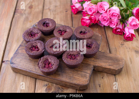 Chocolate Muffins mit Himbeeren und Blaubeeren und weißem Käse Sahne, auf braunem Holz- Board. Ein Strauß frischer Rosen für Liebhaber. Hausgemachtes Gebäck zum Frühstück. Freier Platz für Text Stockfoto