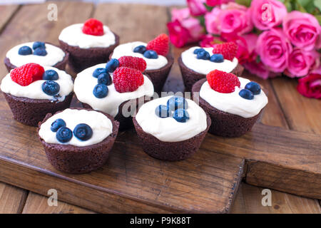 Chocolate Muffins mit Himbeeren und Blaubeeren und weißem Käse Sahne, auf braunem Holz- Board. Ein Strauß frischer Rosen für Liebhaber. Hausgemachtes Gebäck zum Frühstück. Freier Platz für Text. Stockfoto