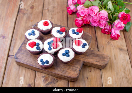 Chocolate Muffins mit Himbeeren und Blaubeeren und weißem Käse Sahne, auf braunem Holz- Board. Ein Strauß frischer Rosen für Liebhaber. Hausgemachtes Gebäck zum Frühstück. Freier Platz für Text. Stockfoto