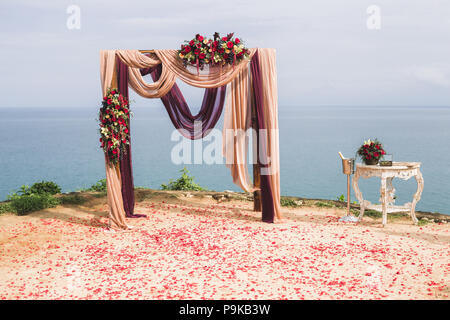 Trauung in Boho Stil mit Meerblick. Mit rote Rosen, gelbe Orchideen, Traube und Lila und beige Stoff Stockfoto