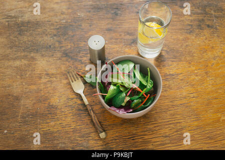 Schale mit frischem Salat Mangold und Spinat Blätter mit Glas Wasser mit Zitrone auf hölzernen Tisch Stockfoto
