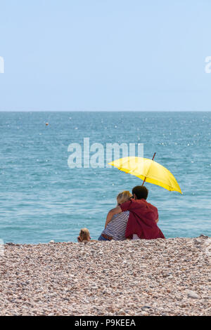 In Worthing, Großbritannien; 14. Juli 2018; Rückansicht Paar am Strand unter einem gelben Regenschirm im heißen Sommer Wetter Stockfoto