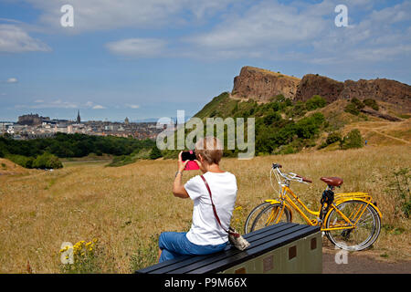 Edinburgh, Schottland, Großbritannien. 19. Juli 2018, Wetter in Großbritannien, Edinburgh Holyrood Park, Schottland, mit Schloss und Stadt im Hintergrund nimmt Frau Handyfoto auf und genießt 20 Grad sonniges Wetter. Stockfoto