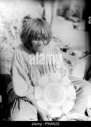 . Englisch: Apache Indian Frau mit einem Korb, ca. 1900 Foto einer Apache Indian Frau mit einem Korb, ca. 1900. Sie sitzt crosslegged auf dem Boden mit einer teilweise bearbeiteten flachen Korb in die Hände in ihren Schoß. Teil eines großen Korb sichtbar ist neben ihr sitzen. Sie trägt ein Plaid Shirt über ein gestreiftes Kleid. Ihr Haar über die Schultern abgeschnitten und hat Bangs. Pinsel wächst hinter ihr. Rufnummer: CHS-3582 Fotograf: James, George Wharton Dateiname: CHS-3582 Abdeckung Datum: ca. 1900 Teil der Sammlung: California Historical Society Collection, 1860-1960 Typ: Bilder Pa Stockfoto