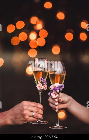 Zwei Gläser Champagner in Paar Hände mit frischen Blumen und Licht orange Blendung auf Hintergrund Stockfoto