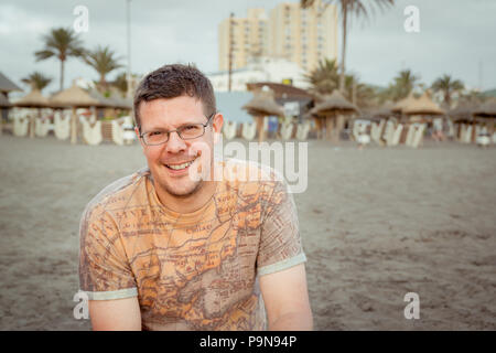 Porträt einer kaukasischen Mann in den Dreißigern oder Vierzigern mit braunen Haaren, Brille, Stoppeln trägt ein T-Shirt Vintage Karte an einem einsamen Strand am Abend Stockfoto