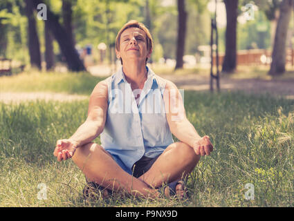 Yoga im Park. Ältere Frau in einem meditativen Yoga pose sitzen auf grünem Gras. Konzept der Ruhe und Meditation. Stockfoto