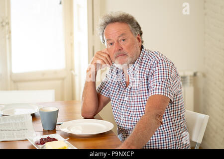 Portrait von depressiven älteren alten Mann mit grauem Haar lesen die Zeitung beim Frühstück und trinken einen Kaffee im Alter in den Ruhestand Konzept. Stockfoto